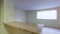 This single-floor space features an open-concept layout that connects the kitchen and living areas seamlessly. A prominent breakfast bar with a granite-patterned laminate countertop serves as the dividing element between the two zones, offering both a casual dining surface and a functional workspace. The countertop's warm beige and gold tones add a subtle visual anchor to the otherwise neutral interior.

The living area is finished with light-colored carpeting, contributing to a clean and understated aesthetic. The walls are painted in a soft, neutral tone throughout, creating a cohesive and versatile backdrop suitable for a variety of furnishing styles. A window on the far wall allows natural light to filter into the living space, brightening the room and providing ventilation. The window is fitted with horizontal blinds, offering adjustable light control and privacy.

The ceilings are smooth and painted white, maintaining a simple, uncluttered appearance. Overall, the space presents a functional and practical layout with clean finishes, making it well-suited for everyday living.