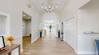 Located at 2351 Pine Brook Dr in Kissimmee, FL, this thoughtfully designed single-floor space impresses with its grand architectural character and bright, welcoming atmosphere. The sweeping entryway sets the tone immediately, featuring soaring vaulted ceilings, a statement chandelier, warm light wood flooring, and a dramatic arched transom window above the front French doors that floods the space with natural light.

The open-concept main area seamlessly integrates the living room, dining room, and kitchen. The living room is anchored by an elegant tile-surround fireplace framed by detailed wainscoting panels, large windows with outdoor views, and stylish pendant lighting. The kitchen features white cabinetry, a mosaic tile backsplash, a stone-topped peninsula, and pass-through views to the entry hallway. The adjacent dining area benefits from floor-to-ceiling windows and access doors that invite the outdoors in.

Throughout the space, consistent wainscoting, recessed lighting, and light wood flooring create a cohesive, refined aesthetic. A private office provides a quiet, functional retreat with its own window and natural light. Two hallways connect the layout efficiently, ensuring smooth flow between areas. The overall ambiance is bright, airy, and polished—ideal for both everyday use and welcoming guests.