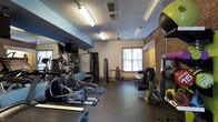 This fitness room offers a comprehensive workout environment designed to accommodate a variety of exercise routines. The space is characterized by an exposed brick accent wall that adds a classic, industrial aesthetic, complemented by white painted walls and a blue accent ceiling border. Rubber flooring throughout the room provides durability and safety underfoot, ideal for high-intensity workouts.

The cardio section is well-stocked with elliptical trainers and a rowing machine, while wall-mounted flat-screen TVs positioned above a large mirror allow users to monitor their form or stay entertained during workouts. The strength-training area includes cable machines, a multi-function press station, and a dedicated bench, catering to both beginners and experienced athletes.

Natural light filters in through windows fitted with horizontal blinds, enhancing the bright, energetic atmosphere created by the fluorescent overhead lighting and a ceiling fan that aids air circulation. A rack along the brick wall holds medicine balls and resistance accessories, ensuring all essential fitness tools are readily accessible within this thoughtfully organized space.