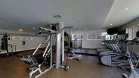 This fitness center is a fully equipped, single-level space designed to accommodate a wide variety of workout routines. The layout is thoughtfully organized, dividing the area into distinct strength training and cardiovascular zones. On the strength side, a multi-station cable machine serves as the centerpiece, accompanied by additional resistance equipment and free weight stations, allowing users to perform a broad range of exercises. Rubberized flooring in the strength area provides durability and traction underfoot. The cardio section, positioned along one wall, features a row of treadmills and elliptical trainers, ideal for sustained aerobic training. Mirrored walls enhance the sense of space and allow users to monitor their form during workouts. Recessed lighting throughout the ceiling provides consistent, even illumination across the entire facility. Windows along one wall allow for natural light to supplement the interior lighting. The neutral color palette of the walls and flooring gives the space a clean, functional aesthetic. Overall, the fitness center is practical and well-appointed, offering residents or members the equipment needed for a comprehensive exercise regimen.