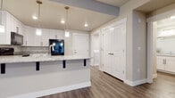 This well-appointed single-floor unit showcases a thoughtfully designed open-concept layout that connects the kitchen and living areas seamlessly. The kitchen is a standout feature, equipped with crisp white shaker-style cabinetry with matte black hardware, granite stone countertops, and a suite of black Whirlpool appliances including a range, over-the-range microwave, and refrigerator. A peninsula with bar-height seating and elegant pendant lighting overhead adds both functionality and visual appeal, making it an ideal space for casual dining or entertaining. The flooring throughout the main living areas consists of warm-toned hardwood-style planks that lend a clean, contemporary feel. The bedroom offers plush carpeting and a bright window with blinds, providing a comfortable and quiet retreat. An in-unit laundry area is conveniently accessible from the main corridor, adding everyday practicality. The bathroom is finished with matching granite countertops, white cabinetry, matte black hardware, and wood-style flooring, maintaining a cohesive aesthetic throughout. Neutral wall tones, recessed lighting, and white panel doors contribute to a bright, modern ambiance across the entire unit.