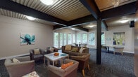 The Schoettler Village Club House in Chesterfield, Missouri is a thoughtfully designed single-story community facility serving residents of the Schoettler Village property. The building's interior is defined by a distinctive corrugated metal ceiling supported by exposed steel beams, lending an industrial-modern aesthetic throughout the space.

Guests enter through glass double doors into a spacious foyer with wood-look flooring, which transitions seamlessly into the main areas of the clubhouse. A warm, wood-paneled reception desk serves as the central hub, where staff can assist residents and visitors. Informational displays highlight community amenities and available packages.

Adjacent to the entry, a dedicated leasing office features two workstations, patterned carpet, and a window allowing natural light to filter in. The main lounge area is generously proportioned, furnished with comfortable sofas and accent chairs arranged around coffee tables, ideal for socializing or relaxing. A wall-mounted flat-screen television adds to the comfort of this gathering space.

Large windows and glass doors along the perimeter of the lounge frame views of an outdoor deck and surrounding greenery, filling the interior with natural light and creating a connection to the outdoors. Smaller seating nooks with round tables provide quieter spaces for conversation or individual use. The overall ambiance is casual yet polished, blending practicality with a welcoming community atmosphere.
