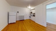 This single-floor unit offers an efficient and well-organized layout comprising a living room, hallway, and full bathroom. The living area is characterized by warm, honey-toned hardwood floors that run throughout, crisp white walls, and a window that admits natural light and outside views, lending the space a bright and open feel despite its compact footprint. A kitchenette is seamlessly integrated into the main living area, featuring white cabinetry, a white electric range, a refrigerator, a classic cast-iron radiator, and a tiled backsplash that adds a subtle decorative touch. The entry door is conveniently positioned adjacent to the kitchen, creating a natural transition into the living space. The bathroom is finished with floor-to-ceiling neutral-toned square tiles, a white soaking tub with an overhead shower, and tile flooring that complements the overall clean aesthetic. A flush ceiling light fixture provides consistent illumination throughout the main area. The unit's layout is straightforward and functional, making efficient use of every square foot. The consistent use of white finishes, natural wood flooring, and understated fixtures gives the space a cohesive and tidy character throughout.
