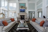 This well-appointed single-floor community space is designed for both comfort and functionality, showcasing an open, flowing layout that connects multiple distinct areas seamlessly. Soaring double-height ceilings define the interior, creating an airy, grand atmosphere throughout. Light wood-look flooring runs continuously across the space, complementing the soft sage and white color palette on the walls and architectural millwork.

The central lounge area serves as the heart of the space, anchored by a stately fireplace with a detailed white surround, stone tile hearth, and a mounted television above the mantel. Generous seating arrangements and layered area rugs contribute to an inviting, relaxed atmosphere. Floor-to-ceiling windows flood the space with natural light and offer views of the surrounding exterior.

Adjacent to the lounge, a dining area features a large table with patterned upholstered chairs, complemented by pendant lighting overhead. Bold red leather accent chairs add a vibrant contrast to the neutral tones. A sleek bar counter with woven leather barstools connects to a fully equipped kitchen, which features white cabinetry, a mosaic tile backsplash, dark granite countertops, and built-in shelving. French doors and transom windows further enhance the bright, open character of this refined and versatile gathering space.