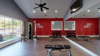 This fitness center in McDonough, GA is a thoughtfully designed, single-floor facility divided into two distinct workout zones, each tailored to a specific training focus. The strength training area commands attention with its bold red accent walls, vaulted ceilings, and a ceiling fan that keeps the space comfortable during intense sessions. Large wall-mounted mirrors amplify the sense of space and allow for proper form checks, while motivational wall decals—including the phrase "I Can and I Will"—set an energizing tone. This zone is outfitted with a cable machine, multiple sets of dumbbells on tiered racks, adjustable weight benches, and a medicine ball rack featuring an assortment of stability balls. Durable hardwood-style flooring runs throughout this area, lending a clean, polished aesthetic. The adjoining cardio zone transitions to textured carpet tiles and a cooler gray-and-red color palette. Motivational phrases such as "Stronger Than Yesterday" and "Determination" adorn the walls, reinforcing the facility's encouraging atmosphere. This section houses treadmills, elliptical trainers, and stationary cycling bikes, with natural light streaming through windows that offer views of surrounding greenery. A wall-mounted sanitization station and waste receptacle reflect thoughtful attention to cleanliness and user convenience. Together, both zones create a comprehensive, motivating environment for residents seeking a full-spectrum fitness experience.