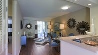 This single-floor residence showcases an open-concept layout that seamlessly connects the kitchen, dining, and living areas into one cohesive and functional space. The kitchen features a light-colored countertop that extends outward as a breakfast bar, offering a natural transition into the combined dining and living zone.

The living area is finished with rich, dark hardwood-style flooring that runs throughout, complemented by neutral-toned walls accented with warm earth tones. A large area rug with a bold chevron pattern in teal and brown anchors the seating arrangement, which centers around a dark sofa and a glass-topped coffee table.

The dining area sits adjacent to the living space and is furnished with upholstered blue chairs, adding a vibrant contrast to the overall color palette. Decorative sunburst mirrors mounted on the walls contribute to a stylish, contemporary aesthetic.

Large sliding glass doors with vertical blinds and striped drapes allow abundant natural light to fill the space while providing access to an outdoor balcony, offering views of the surrounding greenery. Overhead flush-mount lighting ensures the space remains bright and well-lit throughout.