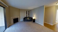 This single-floor space centers around a living room that blends comfort and simplicity. The room features a tiled fireplace along one wall, providing a functional focal point and a natural gathering area. Walls are painted in a warm tan and cool grey palette, creating a subtle contrast that adds visual interest while maintaining a calm, neutral atmosphere. The space is finished with plush wall-to-wall carpeting that contributes to a comfortable, relaxed feel underfoot, while a section of hardwood flooring is visible at the room's edge, hinting at additional areas beyond. A textured popcorn ceiling adds a classic touch overhead. The room is lit by a combination of ambient lighting and a floor lamp positioned near a dark sofa, which serves as the primary seating area. A bold piece of vertical artwork adorns the back wall, providing a striking decorative accent. A baseboard heating unit runs along the lower wall, indicating a practical heating solution throughout the space. The overall layout is open and uncluttered, offering a versatile living area that can accommodate various furniture arrangements and uses.