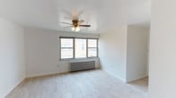 This single-floor space offers a clean, minimalist layout designed for comfortable living. The main living area is finished with light-toned tile flooring that reflects natural light and lends an open, airy feel to the room. Crisp white walls throughout create a neutral backdrop that suits a variety of interior styles.

A multi-pane window positioned along one wall serves as a key architectural feature, drawing in generous amounts of natural light while offering views of the surrounding exterior. Beneath the window sits a classic cast-iron radiator, providing an efficient and traditional heating solution that adds a touch of character to the otherwise contemporary space.

A ceiling fan with an integrated light fixture is centrally mounted overhead, contributing both to air circulation and ambient lighting. The clean lines of the baseboards and the smooth ceiling finish reflect a well-maintained interior.

The overall ambiance of the space is light, open, and functional — ideal for those seeking a straightforward layout with practical features. The combination of natural light, neutral finishes, and classic heating elements gives this space a balanced character that is both inviting and versatile.
