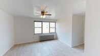This single-floor space offers a clean, minimalist layout designed for comfortable living. The main living area is finished with light-toned tile flooring that reflects natural light and lends an open, airy feel to the room. Crisp white walls throughout create a neutral backdrop that suits a variety of interior styles.

A multi-pane window positioned along one wall serves as a key architectural feature, drawing in generous amounts of natural light while offering views of the surrounding exterior. Beneath the window sits a classic cast-iron radiator, providing an efficient and traditional heating solution that adds a touch of character to the otherwise contemporary space.

A ceiling fan with an integrated light fixture is centrally mounted overhead, contributing both to air circulation and ambient lighting. The clean lines of the baseboards and the smooth ceiling finish reflect a well-maintained interior.

The overall ambiance of the space is light, open, and functional — ideal for those seeking a straightforward layout with practical features. The combination of natural light, neutral finishes, and classic heating elements gives this space a balanced character that is both inviting and versatile.
