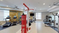 This fitness center offers a comprehensive and energizing environment for exercise enthusiasts of all levels. The space is anchored by a striking yellow motivational mural that covers an entire wall, displaying fitness-themed words and phrases that set an inspiring tone throughout the room. Recessed lighting and ceiling fans maintain a bright and comfortable atmosphere, while large windows along one wall allow natural light to fill the space.

The facility is outfitted with a diverse range of equipment to support varied workout routines. A prominent red boxing station with a heavy punching bag serves as a central feature, ideal for high-intensity training. Cardio equipment includes treadmills and recumbent bikes, positioned to allow ample space between machines. A wall-mounted flat-screen television provides entertainment during workouts.

The flooring is light-toned and easy to maintain, complementing the clean, modern aesthetic of the room. The layout is thoughtfully arranged to maximize usable floor space while ensuring comfortable movement between stations. Overall, this fitness center combines functional equipment, motivating design elements, and a well-organized layout to create an inviting space for daily exercise and wellness routines.