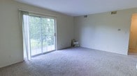 This single-floor space offers a clean, functional layout designed with comfort and simplicity in mind. The main living area is a standout feature, showcasing soft, plush wall-to-wall carpeting that adds warmth and a welcoming feel underfoot. Neutral, light-toned walls throughout the space create a calm and versatile backdrop, making it easy to personalize with any décor style. A large sliding glass door serves as both a striking architectural element and a practical feature, flooding the room with an abundance of natural light while providing direct access to the outdoors. The view through the glass door reveals a lush, green tree canopy, bringing a refreshing sense of nature into the interior. Overhead, a smooth white ceiling with a visible HVAC vent speaks to the thoughtful functionality built into the design. The overall ambiance is bright, airy, and move-in ready, with an open feel that maximizes the sense of space. Whether used as a living or leisure area, this space offers a tranquil setting enhanced by its strong connection to the natural surroundings visible just beyond the glass.