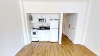 This single-floor unit offers a clean, minimalist aesthetic defined by bright white walls and warm hardwood flooring that runs throughout the open layout. The kitchenette is neatly tucked into a recessed alcove, maximizing the sense of open space in the main living area. It is outfitted with white shaker-style cabinetry providing ample storage, a dark countertop with an integrated sink, a freestanding electric range, a full-size refrigerator, and a built-in microwave overhead. An electrical panel is conveniently accessible within the kitchen alcove as well. The open floor plan allows the kitchenette to flow seamlessly into the rest of the living space, creating an airy and uncluttered feel. A doorway visible from the main area leads to an additional room or hallway, suggesting further functional space beyond the primary living zone. The entry door is equipped with a deadbolt, adding a practical security feature. The overall ambiance is bright, clean, and efficient — well-suited for comfortable everyday living. The consistent use of white tones paired with natural wood flooring lends the space a fresh, modern character.