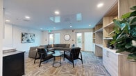 Located at 7288 Maple Street in Mission, BC, this thoughtfully designed communal space offers a welcoming and functional environment across a single floor. The layout is divided into two distinct areas, each serving a unique purpose while maintaining a cohesive contemporary aesthetic throughout.

The main lounge area is anchored by a generous sectional sofa and complemented by sleek leather accent chairs arranged around a minimalist coffee table — ideal for relaxed gatherings or casual meetings. Patterned carpet tiles add texture underfoot, while recessed lighting and large exterior doors allow natural light to fill the room. A built-in wall unit with warm wood-toned shelving houses a flat-screen TV and decorative accessories, adding both function and visual warmth. A compact kitchenette with cabinetry, shelving, and a microwave is integrated seamlessly along one wall, enhancing the space's utility.

The second area functions as a private meeting room, enclosed by sleek black-framed glass partitions that maintain visual openness while providing separation. A large oval marble-top table surrounded by navy upholstered chairs serves as the focal point, with abstract artwork adorning the clean white walls. The overall ambiance is modern and professional, balancing comfort with practicality in a polished, well-lit setting.