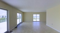 This single-floor space offers a clean, open layout bathed in natural light, with a design that emphasizes the stunning coastal surroundings. The main living area is generously proportioned, featuring large-format light tile flooring that extends throughout, lending a sense of continuity and spaciousness to the interior.

Two sets of sliding glass doors dominate the exterior wall, framing panoramic ocean views and allowing abundant natural light to pour into the space. These doors provide direct access to an outdoor area, seamlessly connecting the interior to the coastal environment beyond. Palm trees and the open water are visible through the glass, reinforcing the beachside character of the space.

The walls are finished in a soft, warm neutral tone that complements the bright, airy atmosphere, while the textured white ceiling adds a subtle architectural detail. The overall design is straightforward and unfussy, prioritizing the views and natural light as the primary features. The open floor plan allows for flexible use of the space and creates a flowing, unobstructed layout throughout. This space is well-suited for those seeking a coastal living environment defined by light, openness, and direct visual connection to the ocean.