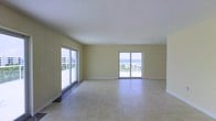 This single-floor space offers a clean, open layout bathed in natural light, with a design that emphasizes the stunning coastal surroundings. The main living area is generously proportioned, featuring large-format light tile flooring that extends throughout, lending a sense of continuity and spaciousness to the interior.

Two sets of sliding glass doors dominate the exterior wall, framing panoramic ocean views and allowing abundant natural light to pour into the space. These doors provide direct access to an outdoor area, seamlessly connecting the interior to the coastal environment beyond. Palm trees and the open water are visible through the glass, reinforcing the beachside character of the space.

The walls are finished in a soft, warm neutral tone that complements the bright, airy atmosphere, while the textured white ceiling adds a subtle architectural detail. The overall design is straightforward and unfussy, prioritizing the views and natural light as the primary features. The open floor plan allows for flexible use of the space and creates a flowing, unobstructed layout throughout. This space is well-suited for those seeking a coastal living environment defined by light, openness, and direct visual connection to the ocean.