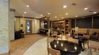 This inviting single-floor dining and lounge space combines functionality with a warm, hospitable ambiance. The open-concept layout seamlessly connects a bar and counter seating area with a spacious dining room, offering a variety of seating arrangements to accommodate different group sizes. Dark wood round tables paired with light wood chairs provide a balanced contrast, while a patterned carpet defines the lounge zone alongside clean tile flooring in the main dining area.

The bar area is well-appointed with wooden cabinetry, open shelving stocked with glassware and decorative accents, and a tile backsplash that adds texture and visual interest. Pendant lighting above the bar counter complements the recessed ceiling lights throughout, casting a warm, even glow across the space.

Sturdy stone or brick-clad columns anchor the room architecturally, adding a rustic yet refined character to the interior. The vaulted ceiling in portions of the space enhances the sense of openness. Neutral-toned walls and warm wood finishes across cabinetry and trim elements create a cohesive, comfortable atmosphere. A glass-paned door allows natural light to filter in, connecting the interior to an adjacent area. Overall, the space is well-designed for communal dining and socializing.