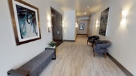 This single-floor space presents a thoughtfully designed corridor that combines functionality with upscale aesthetics. The hallway features striking marble-style flooring with warm beige and grey veining tones that run throughout, lending an air of elegance to the space. Neutral white walls provide a clean backdrop, accented by sleek wall-mounted sconce lighting that casts a warm, ambient glow along the length of the corridor.

Carefully selected furnishings enhance the space's refined character. A low-profile grey upholstered bench sits against one wall, accompanied by a minimalist side table topped with a small decorative plant. On the opposite end, a pair of charcoal-toned armchairs with mid-century modern styling offer a comfortable seating arrangement. Large framed artwork punctuates the walls — including an abstract blue and green piece and a dramatic dark marbled composition — adding visual interest and a gallery-like quality. Dark espresso-toned double doors with modern hardware serve as a prominent architectural feature. The overall ambiance is professional yet inviting, suggesting a high-end commercial or hospitality environment.