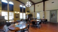 This single-floor space presents an impressive dining or community gathering area defined by its dramatic vaulted ceilings accented with dark exposed wood beams. A classic chandelier hangs as a central focal point, casting warm light across the room and complementing the rich tones of the polished hardwood flooring. The walls are finished in warm golden-yellow hues, contributing to an inviting and comfortable atmosphere.

Tall arched windows along the upper walls and standard windows along the lower walls flood the interior with natural light, enhancing the sense of openness and volume. The space is furnished with multiple round dining tables paired with sturdy chairs, creating a layout well-suited for communal dining or social gatherings. Tables are accented with small floral centerpieces and decorative touches including small American flags, adding a personable character to the room.

A brick accent wall element and framed artwork add architectural interest, while a flat-screen television is mounted at a mid-height position for convenient viewing. A beverage station with a Coca-Cola cooler is visible along one wall, suggesting a functional hospitality setup. Overall, the space balances a warm, homey aesthetic with practical amenities.