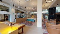 Located at 1777 Wewatta Street in Denver's Union Station neighborhood, the Grand Hall is a expansive, single-floor amenity space designed with a contemporary aesthetic that blends warm wood tones, industrial accents, and curated furnishings throughout. The space is introduced by a light-filled entry foyer with soaring double-height ceilings, setting an immediately welcoming tone.

The main gathering area features multiple distinct lounge zones, including seating arrangements with teal sofas, leather chairs, and patterned area rugs, as well as a large communal wooden work table surrounded by black chairs beneath geometric hexagonal pendant lighting. A dramatic wood-clad staircase with integrated LED step lighting connects to an upper mezzanine level, complemented by a striking black spiral staircase in a secondary lounge area.

The full chef's kitchen showcases a large marble-topped island, professional appliances, copper fixtures, and a bold mural as a focal point. A game room with a billiard table and long bar-height communal tables adds a recreational dimension. Floor-to-ceiling glass doors open directly onto an outdoor patio terrace with views of the surrounding Denver streetscape, flooding the interior with natural light.