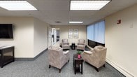 This single-floor commercial space is thoughtfully designed to support both comfort and functionality across several distinct areas. The lounge areas are furnished with plush, neutral-toned armchairs arranged around dark side tables, creating a welcoming and relaxed atmosphere. Decorative framed artwork adorns the walls, and patterned carpet flooring ties the space together with a professional yet inviting aesthetic. Large windows fitted with vertical blinds allow natural light to filter in while maintaining privacy, offering views of the surrounding greenery outside.

The kitchen is equipped with rich, dark wood cabinetry, a double-basin sink, and full-sized appliances including a refrigerator, electric range, microwave, and dishwasher. Light-toned countertops extend into a generous bar-style surface, making the kitchen practical for meal preparation and service. Tile flooring throughout the kitchen area ensures durability and ease of maintenance.

Adjacent to the kitchen is a spacious communal dining or meeting room featuring multiple round tables with ergonomic seating for groups. Recessed fluorescent lighting and large windows with vertical blinds keep the room bright and airy. A colorful piece of wall art adds a vibrant accent to the otherwise neutral palette. This versatile space is well-suited for gatherings, training sessions, or collaborative activities.