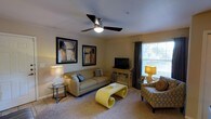This single-floor space offers a thoughtfully designed living area that combines comfort and style. The living room serves as the focal point, featuring warm neutral wall tones complemented by plush carpeted flooring that adds a sense of warmth underfoot. A sleek ceiling fan with an integrated light fixture is centrally mounted, providing both air circulation and ambient lighting throughout the room. Natural light streams in through a large window dressed with elegant curtain panels, offering a pleasant view of the surrounding greenery outside. The space is furnished with a generously sized sofa adorned with decorative accent pillows, paired with a patterned armchair that adds visual interest. A distinctive yellow sculptural coffee table serves as a bold centerpiece, injecting a vibrant pop of color against the otherwise subdued palette. The entertainment area features a flat-screen television atop a wooden media console, while framed abstract artwork adorns the walls, contributing to the overall modern aesthetic. Accent table lamps on either side of the room provide layered lighting options, creating a warm and inviting ambiance. The layout is open and well-organized, making efficient use of the available space while maintaining a comfortable flow throughout.
