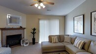 This single-story space presents a well-appointed living area designed for comfort and functionality. The living room serves as the central feature, anchored by a large sliding glass door dressed with vertical blinds that allow for adjustable natural light control. The ceiling is finished with a textured surface and equipped with a ceiling fan fitted with a light fixture, providing both air circulation and ambient lighting throughout the space.

The flooring is carpeted in a neutral tone, complementing the warm, light-colored walls that give the room an open and inviting feel. A sectional sofa in a muted taupe finish serves as the focal point of the seating area, styled with patterned throw pillows in striped and chevron designs. A low-profile chaise extension adds to the lounging comfort of the arrangement.

To one side of the room, a fireplace with a simple mantle adds an architectural detail that enhances the room's character. Decorative elements including a tall potted plant in a dark planter and ceramic accent pieces contribute to the overall aesthetic. A secondary window on the adjacent wall provides additional natural light, further brightening the space.