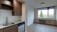 This single-floor unit offers a thoughtfully designed open-concept layout that efficiently combines kitchen and living spaces. The kitchen features rich, dark wood cabinetry with sleek hardware, light-toned countertops, a built-in sink, and a dishwasher, all complemented by a clean white subway tile backsplash that adds a contemporary touch. The cabinetry extends vertically, maximizing storage within the compact footprint.

The living area flows naturally from the kitchen, unified by continuous light wood laminate flooring that lends warmth and cohesion throughout. A standout feature of the living space is a large picture window that frames an inviting view of mature evergreen trees and surrounding scenery, filling the room with natural light. Beneath the window, a built-in ledge provides a functional surface, ideal as a workspace or casual seating area. A wall-mounted HVAC unit ensures year-round climate control.

The clean white walls and minimalist design approach create an airy, uncluttered atmosphere. A doorway visible from the main space suggests access to a separate room or bathroom, adding functional versatility to the unit's overall layout.