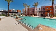 Located at 1115 E Florence Blvd in Casa Grande, Arizona, this outdoor amenity space is designed to deliver a resort-style experience within a contemporary residential community. The centerpiece is a generously sized swimming pool with crystal-clear water, framed by a spacious sun deck finished in aggregate concrete — ideal for lounging under the Arizona sun. Tall, mature palm trees are strategically positioned throughout the area, providing both shade and a striking visual presence against the backdrop of the vibrant desert sky.

The surrounding deck is well-appointed with an array of chaise lounges, shaded umbrella seating, and cozy cabana-style pod chairs, offering a variety of relaxation options. Decorative oversized planters in bold blue and terracotta tones add colorful accents throughout the space.

Thoughtfully designed privacy screens with geometric patterning define intimate seating nooks and garden areas, blending aesthetic appeal with functional separation. Native and drought-tolerant plantings complement the desert setting. The multi-story buildings surrounding the courtyard feature a modern architectural palette of white, orange, and neutral tones, with private balconies overlooking the amenity space. The overall ambiance is vibrant, welcoming, and well-suited to the warm Arizona climate.