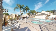 This outdoor community amenity space offers a well-appointed recreational area centered around a generously sized swimming pool with clear, blue water and convenient step entry points. Adjacent to the main pool sits a circular spa, providing a relaxing complement to the larger swimming area. The pool deck is finished in concrete with accent tile borders and is furnished with multiple white lounge chairs, offering ample space for sunbathing and leisure. The area is enclosed by a combination of metal fencing and masonry block walls, with gated access points secured by latch hardware and exterior lighting fixtures mounted above entry points. Tall, mature palm trees are planted throughout the space, lending a tropical, resort-like atmosphere while providing partial shade. Decorative stone seating and tables add additional gathering options near the pool. A separately enclosed tennis court with a reddish surface and chain-link fencing is also part of the complex, providing an additional active recreation option. The surrounding buildings feature stucco exteriors and pitched rooflines typical of Southern California residential architecture. Under bright blue skies, the entire outdoor space conveys a sun-drenched, leisure-focused environment suited for year-round enjoyment.
