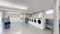 This single-floor shared laundry facility is designed for efficiency and practicality, providing a clean and functional environment for laundry needs. The space features a row of commercial-grade front-loading washing machines, numbered for easy identification, lined up along one wall and elevated on a platform for convenient access. Complementing these are top-loading dryers positioned along the adjacent wall, offering a full laundry solution within one room. The flooring is a smooth, easy-to-clean surface in a neutral gray tone, contributing to the space's hygienic and utilitarian feel. Overhead, fluorescent lighting ensures the room is brightly and evenly lit throughout. The walls are finished with a two-tone treatment — a lighter upper section and a blue lower panel — adding a modest touch of character to the otherwise functional space. Exposed ductwork and ventilation pipes run along the ceiling and walls, indicative of the space's practical, no-frills design. A laundry cart is available for user convenience. A small window allows for a degree of natural light, complementing the overhead fixtures. Overall, this laundry facility is well-maintained and equipped to serve a high volume of use efficiently.