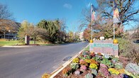 The LeSilve community presents a welcoming exterior environment characterized by thoughtfully maintained grounds and classic architectural elements. At the entrance, a brick monument sign is flanked by vibrant seasonal flower beds featuring chrysanthemums and ornamental plantings in warm autumn hues of yellow, orange, and purple, accented by decorative pumpkins atop the signage structure. A flagpole stands prominently at the entry drive.

The surrounding landscape showcases a mix of mature deciduous and evergreen trees displaying rich fall foliage under open blue skies. A smooth, curving concrete pathway winds through well-kept green lawns, guiding residents and visitors through the grounds. Traditional-style lamp posts line the walkway, contributing to the community's classic aesthetic.

The multi-story residential building visible in the background features a neutral stone or brick exterior with pitched rooflines and balconies. The grounds include open grassy areas and what appears to be a water feature or pond nearby, enhancing the natural ambiance. Overall, the LeSilve community conveys a well-established, park-like setting with attention to landscaping detail and a tranquil outdoor atmosphere.