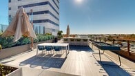 Located at 18750 Delaware St in Huntington Beach, California, this rooftop terrace offers an expansive open-air amenity space designed for both relaxation and social gatherings. The deck is finished with light wood-toned porcelain tile flooring that lends a warm, contemporary aesthetic throughout.

The terrace is thoughtfully organized into distinct functional zones. A dining area features a long white rectangular table surrounded by stylish blue woven chairs, ideal for group meals or casual meetings. Adjacent to this, a built-in outdoor kitchen includes a full-size gas grill, counter space, and a utility sink, providing everything needed for outdoor cooking and entertaining.

A bar counter with green upholstered barstools serves as a social hub, while woven egg-shaped lounge chairs and cushioned seating areas create comfortable spots for relaxation. A foosball table adds a recreational element to the space. Lush raised planters with tropical and Mediterranean greenery are distributed throughout, providing natural privacy screening and vibrant color.

The terrace is enclosed by glass railing panels that preserve unobstructed panoramic views of the surrounding Huntington Beach cityscape, including palm trees and the horizon under open Southern California skies. The abundance of natural sunlight makes this rooftop a bright, inviting destination throughout the day.