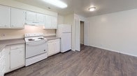 This single-floor unit offers a straightforward, functional layout spread across several rooms. The kitchen is well-equipped, featuring white cabinetry extending to the upper walls for generous storage, gray laminate countertops, a double-basin sink, a white electric coil range with overhead ventilation, and a full-size refrigerator. Dark hardwood-style flooring runs throughout the kitchen, complementing the neutral cream-toned walls and providing a practical, easy-to-maintain surface. A louvered closet door near the kitchen offers additional storage access.

The bedrooms are carpeted in a warm beige tone and finished with the same neutral wall palette, creating a calm and cohesive atmosphere. Each room features windows dressed with vertical blinds, allowing natural light to filter in. Several bedrooms include built-in closets, with at least one featuring an open closet with a hanging rod. One bedroom offers a view of mature greenery outside, adding a pleasant natural element.

The bathroom is compact and functional, outfitted with a soaking tub, a toilet, and a vanity with a gray countertop and mirror. The overall design of the unit is clean and understated, with a consistent neutral color scheme throughout that provides a versatile backdrop for a variety of furnishing styles.