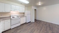 This single-floor unit offers a straightforward, functional layout spread across several rooms. The kitchen is well-equipped, featuring white cabinetry extending to the upper walls for generous storage, gray laminate countertops, a double-basin sink, a white electric coil range with overhead ventilation, and a full-size refrigerator. Dark hardwood-style flooring runs throughout the kitchen, complementing the neutral cream-toned walls and providing a practical, easy-to-maintain surface. A louvered closet door near the kitchen offers additional storage access.

The bedrooms are carpeted in a warm beige tone and finished with the same neutral wall palette, creating a calm and cohesive atmosphere. Each room features windows dressed with vertical blinds, allowing natural light to filter in. Several bedrooms include built-in closets, with at least one featuring an open closet with a hanging rod. One bedroom offers a view of mature greenery outside, adding a pleasant natural element.

The bathroom is compact and functional, outfitted with a soaking tub, a toilet, and a vanity with a gray countertop and mirror. The overall design of the unit is clean and understated, with a consistent neutral color scheme throughout that provides a versatile backdrop for a variety of furnishing styles.