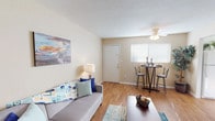 This single-floor, two-bedroom, one-bathroom residence offers a comfortable and functional layout designed to maximize everyday living. The open living area serves as the heart of the home, featuring warm hardwood-style flooring, neutral beige walls, and a ceiling fan that contributes to a light and airy atmosphere. A wall-mounted flat-screen TV and ample space for seating make it an inviting area for relaxation. The living space flows naturally toward a dining nook positioned near a large window with blinds, allowing abundant natural light to fill the area. The kitchen is equipped with warm oak cabinetry, granite-style countertops, a double stainless steel sink, and a dishwasher, providing a practical and attractive workspace. A full-size refrigerator completes the kitchen's functionality. The bedrooms feature plush carpeting and windows that allow natural light in, with the primary bedroom offering room for a full suite of furniture. The shared full bathroom includes a clean, white tub-shower combination. Throughout the residence, the consistent use of hardwood-style flooring in common areas and soft carpet in the bedrooms creates a cohesive, comfortable feel well-suited for everyday living.