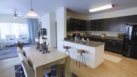 This single-story space showcases an open-concept layout that integrates the kitchen and dining areas into a cohesive, functional living environment. The kitchen is a standout feature, equipped with rich, dark espresso-toned cabinetry that extends to the upper and lower sections of the walls, providing generous storage throughout. A peninsula with a dark granite-style countertop serves as both a prep area and a casual dining spot, accompanied by rustic wooden bar stools that add a warm, organic contrast to the sleek cabinetry. The kitchen is well-appointed with modern appliances, including a built-in microwave, and features a deep basin sink with a contemporary faucet. Overhead, recessed and flush-mount lighting ensures the workspace is well illuminated. The adjacent dining area features a light wood table accented by a striking candelabra centerpiece, complemented by a pendant light overhead. Light hardwood-style flooring runs throughout, tying the spaces together and enhancing the bright, airy atmosphere. The neutral wall tones and open sightlines contribute to a sense of spaciousness, making this layout ideal for both everyday living and entertaining.
