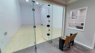 This racquetball court is a purpose-built athletic space designed for both competitive play and recreational use. The playing surface consists of a classic hardwood court floor, clearly marked with the standard boundary lines essential for gameplay. The court's walls are bright white, maximizing visibility and ensuring optimal playing conditions.

A standout architectural feature is the full-height glass panel wall at the court's entrance, constructed with thick, frameless tempered glass secured by sleek black hardware. This transparent boundary allows for unobstructed viewing of the court from the outside while maintaining the integrity of play within.

Adjacent to the glass entry wall is a small spectator area, finished with an elegant herringbone-patterned hard floor. A pair of cushioned gold upholstered chairs are positioned here, providing comfortable seating for observers watching matches. The transition between the spectator zone and the court entrance is clean and well-defined.

Recessed ceiling lighting illuminates the court evenly, ensuring consistent visibility across the entire playing surface. The overall design balances athletic functionality with refined finishes, making this a well-considered facility suitable for players of all skill levels.