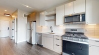 This contemporary single-floor unit showcases a thoughtfully designed open layout with consistent wood-look flooring throughout, creating a cohesive and modern aesthetic. The kitchen is a standout feature, equipped with stainless steel appliances including a gas range, over-the-range microwave, French door refrigerator, and dishwasher. Two-tone cabinetry — white upper cabinets paired with light wood lower cabinets — is complemented by a classic white subway tile backsplash, open floating shelves, and a matte black faucet at the undermount sink. Light-filled and efficient, the kitchen also benefits from proximity to a large window offering natural light. The open living area is bright and airy, featuring recessed lighting, a ceiling fan, and a clean neutral color palette that enhances the sense of space. A central hallway connects the main living zone to the private areas of the unit. The bathroom is finished with a floating wood-toned vanity with drawers and matte black hardware, wood-look flooring, and a walk-in shower enclosed by a glass partition. The bedroom is generously proportioned with wood-look flooring, neutral walls, and direct access to an outdoor balcony, offering views of the surrounding cityscape and an abundance of natural light.