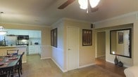 This single-story residence showcases classic architectural detailing throughout its interior. The main living area is finished with elegant crown molding that runs along the ceiling perimeter, adding a refined touch to the space. A ceiling fan with a built-in light fixture is centrally mounted, providing both airflow and ambient lighting to the room.

The living space features neutral-toned carpeting, which transitions to hard flooring in adjacent areas, creating a natural distinction between zones. White painted walls are complemented by white baseboards, contributing to a clean and cohesive aesthetic. Decorative wall art adds character to the space.

An open passageway connects the living area to the kitchen, which is outfitted with white cabinetry offering ample storage. The kitchen includes countertop workspace and standard appliances, maintaining the home's consistent neutral color palette.

Multiple doorways branch off from the main living area, providing access to additional rooms, including what appears to be a hallway leading to further living spaces. The layout offers a functional flow between rooms, making the home practical and easy to navigate throughout.