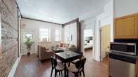 This single-floor residence artfully combines historic architectural character with comfortable, modern living. The open living and dining area serves as the heart of the home, where rich hardwood floors run throughout and striking exposed wooden beams frame the space, adding warmth and industrial charm. Multiple windows allow natural light to fill the room, while a seating arrangement centered around a coffee table creates an inviting atmosphere for relaxation or entertaining.

The kitchen is a standout feature, showcasing a dramatic full exposed brick wall that adds texture and character. It is equipped with stainless steel appliances, ample cabinetry, and a dedicated dining table, making it both functional and visually compelling.

The residence includes multiple bedrooms, each designed with comfort and practicality in mind. Bedrooms feature hardwood flooring, generous windows that bring in natural light, dedicated workspace areas with desks, ample storage with dressers and closets, and radiator heating. The clean, neutral color palette throughout creates a calm and cohesive aesthetic.

The bathroom is neatly appointed with light tile flooring and clean white fixtures. The overall ambiance of the space is one of classic New York character — a thoughtful blend of pre-war architectural details and well-maintained contemporary furnishings.