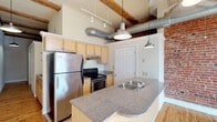 This single-floor space showcases a compelling mix of industrial character and refined detail. A central hallway connects the main areas, lined with hardwood floors and white walls that keep the aesthetic clean and bright. A classic French door with glass panes provides separation between spaces while maintaining an open, airy feel. Pendant barn-style lights hang from the ceiling along the corridor, adding a consistent design thread throughout.

The kitchen is a standout feature, boasting soaring ceilings with exposed wooden beams and visible ductwork that reinforce the industrial loft aesthetic. Maple cabinetry pairs with granite countertops, which extend across a curved kitchen island complete with a double sink—ideal for both prep and casual dining. Stainless steel appliances, including a refrigerator and range, add a modern, functional edge. Exposed brick walls anchor one side of the kitchen, introducing warmth and texture. Natural light filters in from large windows at the far end of the space, complementing the warm tones of the hardwood flooring. The overall ambiance balances raw, warehouse-inspired elements with thoughtful, polished finishes.