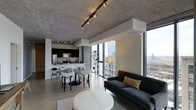 This single-floor unit showcases a seamlessly integrated open-concept layout that combines living, dining, and kitchen areas into one cohesive space. The exposed concrete ceiling adds an industrial character, complemented by a track lighting system that provides flexible, adjustable illumination throughout. Floor-to-ceiling windows line the exterior wall, flooding the interior with abundant natural light while offering sweeping urban skyline views, creating a strong visual connection to the city beyond.

The living area features a charcoal tufted sofa paired with a segmented white ottoman and a patterned area rug, anchoring the lounge space with a contemporary aesthetic. The dining area accommodates a round table with modern white chairs, positioned to take full advantage of the panoramic window views. The kitchen, finished in dark cabinetry with stainless steel appliances, features a breakfast bar with barstool seating and a clean, functional layout. Light wood-tone flooring runs throughout, offering a warm contrast to the cooler concrete and dark finishes. Together, the design elements create a polished, urban-modern ambiance suited for contemporary living.