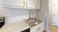 Located at 5 Dorchester Dr in Bethel Park, PA, this single-floor residence has been thoughtfully updated with modern finishes throughout. The kitchen stands out with crisp white cabinetry accented by brushed gold hardware, light granite countertops, a stainless steel sink with a pull-down faucet, and a textured white geometric tile backsplash that adds subtle visual interest. A dishwasher is also included, rounding out the functional kitchen layout. The bathroom features a clean, bright tub-and-shower combination with a square rain showerhead and bronze-tone fixtures, complemented by wood-look tile flooring and neutral walls for a calm, spa-like atmosphere. The living and bedroom areas are finished with light wood-look plank flooring that runs consistently throughout, creating a cohesive and airy feel. Neutral warm-gray wall tones provide a versatile backdrop, while large windows allow natural light to fill the rooms. Baseboard heating is present in the living areas. The open layout, as seen in the 3D model, reveals a well-organized floor plan with clearly defined living, sleeping, kitchen, and bathroom zones, offering practical and comfortable everyday living in a quiet Bethel Park neighborhood.
