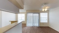 This single-story space features an open-concept layout that connects the kitchen and living areas seamlessly. The kitchen includes overhead cabinetry and a raised countertop bar that serves as a natural divider between the two spaces, offering both functionality and a sense of openness. The living area is carpeted and benefits from abundant natural light streaming through multiple windows fitted with horizontal blinds, as well as a large sliding glass door dressed with vertical blinds that provides outdoor access. A ceiling fan with a light fixture is centrally positioned in the living area, ensuring comfort and additional illumination. The textured popcorn ceilings are consistent throughout, adding a classic residential character to the space. The neutral color palette of beige and white tones across the walls, ceilings, and countertops creates a cohesive and understated aesthetic. Overall, the space offers a practical and comfortable layout with well-distributed natural light and straightforward finishes suited for everyday living.