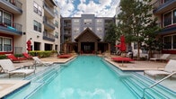 This single-story poolside cabana structure serves as a central amenity hub within a multi-story residential community. Architecturally, the structure features a steeply pitched vaulted ceiling supported by dark-painted wood framing, with recessed lighting and a ceiling fan providing comfort and ambiance throughout the covered area. A large rustic wood plank table anchors the interior, flanked by red upholstered lounge chairs that provide a bold contrast to the neutral gray palette of the horizontal lap siding walls. A wall-mounted television is positioned beneath the peak of the ceiling, making the space functional for outdoor entertainment. The cabana opens directly onto a expansive resort-style swimming pool, offering unobstructed sightlines across the water. The surrounding deck is furnished with additional red chaise lounges, side tables, and picnic-style benches, creating versatile seating options. A pergola-covered entry accented with red brick columns and manicured landscaping provides a welcoming transition point between the interior corridors and the pool courtyard. Semi-enclosed side alcoves offer more intimate seating nooks with built-in red cushioned sofas and individual chairs. The overall ambiance is modern yet inviting, blending industrial-inspired architectural details with vibrant furnishings and open courtyard views.