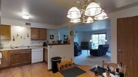 This single-floor residence offers a seamlessly connected living space that combines functionality with a warm, welcoming ambiance. The open-concept design allows the kitchen, dining area, and living room to flow naturally into one another, creating a sense of spaciousness throughout.

The kitchen is equipped with classic wood cabinetry, a white dishwasher, and a raised breakfast bar that serves as both a practical workspace and a casual gathering spot. A wall clock and decorative accents add a homey touch to the area. The kitchen transitions smoothly into the dining space, which features a wooden dining table and chairs beneath a statement brass chandelier with globe-style shades that anchors the room with a classic elegance.

Beyond the dining area, the living room offers a comfortable, carpeted retreat furnished with plush seating and framed by a large sliding glass door that invites generous natural light and provides views of the surrounding outdoor greenery. Warm lamplight complements the natural illumination, creating a cozy atmosphere throughout the day and evening.

The consistent use of warm wood tones in the flooring and furnishings, paired with neutral wall colors, ties the entire space together in a cohesive and inviting aesthetic.