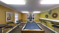 This single-floor recreational space is thoughtfully designed to serve as a social and entertainment hub. The room's warm yellow walls and white crown molding create an inviting atmosphere, complemented by flush fluorescent lighting that ensures the space is well-lit throughout. At the center of the room sits a full-size pool table with a blue felt surface and a classic wooden frame, serving as the focal point of the space. A built-in white shelving unit along the back wall houses an entertainment center, complete with a television, speakers, and display shelving for decorative items, providing an additional entertainment option for guests. Bar-height tables with sleek black stools line both sides of the room, offering ample seating for socializing. A window along one wall allows natural light to filter in, adding brightness to the space. The carpeted flooring provides comfort underfoot and contributes to the room's casual, relaxed ambiance. Overall, this space is well-equipped for group recreation, combining game room functionality with comfortable lounge-style seating in a compact, efficiently arranged layout.