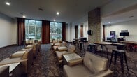 This single-floor amenity space is thoughtfully designed to offer a well-rounded social and recreational experience. The lounge area is a standout feature, furnished with plush leather armchairs and low coffee tables arranged for comfortable conversation. Bold patterned carpeting runs throughout, complemented by floor-to-ceiling windows dressed in striped red and white curtains that allow generous natural light and views of the surrounding greenery. Exposed brick columns add a distinctive architectural character, anchoring the space with a warm, industrial-chic aesthetic.

A dedicated game room extends the social atmosphere further, equipped with a pinball machine, shuffleboard table, poker table, and multiple round dining-height tables with chairs — ideal for casual gatherings and entertainment. Framed artwork adorns the walls, contributing to the curated, hospitality-inspired design throughout.

A corridor connecting the spaces features patterned carpeting and decorative accents, including a framed locomotive print and an ornamental console, lending a polished transitional area between zones. Glass-paneled doors offer a clear view into an adjoining fitness center stocked with exercise equipment. Recessed lighting throughout ensures a consistently well-lit environment, while the cohesive design palette of neutral tones, brick, and bold accents ties the entire space together seamlessly.