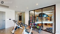 This expansive fitness center delivers a thoughtfully designed workout environment across a single, generously proportioned floor. The space is organized into distinct functional zones, seamlessly flowing from one to the next while maintaining an open, airy feel throughout.

The main exercise floor features durable light-toned flooring and is illuminated by recessed ceiling lighting, ensuring a bright and energizing atmosphere. A striking floor-to-ceiling panoramic sunset mural serves as a dramatic focal point, adding warmth and visual depth to the space. The free weights area is well-equipped with a full dumbbell rack, adjustable benches, medicine balls, and plyo boxes to accommodate a wide range of strength and conditioning workouts.

Multiple cable machines and strength training stations are positioned throughout, supported by structural columns that subtly define training areas without interrupting the open layout. An upper-level mezzanine, accessible via a staircase with sleek cable railings, features a slatted wood ceiling detail and large pendant lighting, offering additional equipment space with an elevated industrial-chic aesthetic.

Exposed brick walls add a raw, textural contrast to the clean white ceilings and light hardwood flooring in transitional areas. Floor-to-ceiling windows along one side of the space flood the interior with natural light and provide views to the outdoors, enhancing the overall sense of openness and vitality.