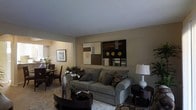 This single-floor residence offers a thoughtfully designed interior with a warm, inviting ambiance carried throughout its open living spaces. The living room serves as the centerpiece of the home, featuring a plush, gray upholstered sofa adorned with decorative throw pillows, anchored by a dark, rounded coffee table and a soft area rug. A pair of bold contemporary art panels in earth tones — brown, black, and gold — commands attention on the main wall, adding a stylish focal point to the room. A classic white table lamp atop a dark side table provides both function and elegance. Adjacent to the living area, the dining space accommodates a small table and chairs beneath warm lighting, with lush greenery adding a natural, refreshing touch. The walls throughout are finished in a soft, neutral beige tone that complements the overall palette and creates a sense of warmth. The textured white ceiling adds subtle architectural interest. Natural and artificial lighting work in harmony to keep the space feeling bright and welcoming. The overall design reflects a tasteful blend of comfort and contemporary style, making it a functional and aesthetically pleasing living environment.